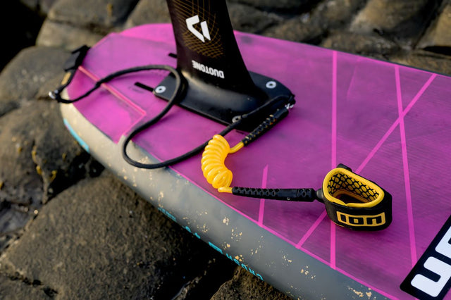 ION Wing Board Leash Core Hybrid Ankle 2026, schwarzes Neopren mit gelbem Kunststoffverschluss, auf magentaboard liegend
