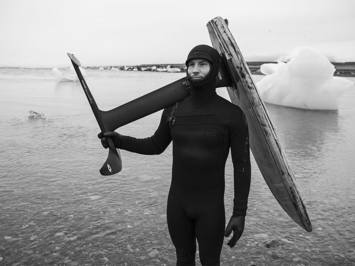 Surfer in schwarzem Neoprenanzug trägt Surfboard am Strand, Schwarz-Weiß-Fotografie
