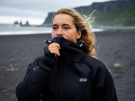 ION Neo Shelter Jacket 2024 schwarz, Frau trägt Jacke mit hochgezogenem Kragen am Strand, Frontalansicht