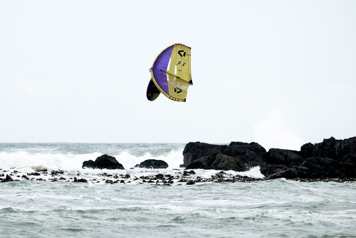 ION Wing / SUP Leash Core Coiled Hip 2024, gelb-violette Kitewing über Meer mit Felsen, Seitenansicht