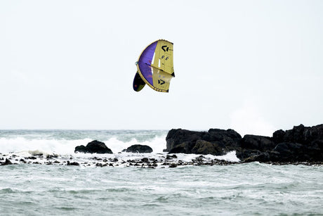 ION Wing / SUP Leash Core Coiled Hip 2024, gelb-violette Kitewing über Meer mit Felsen, Seitenansicht