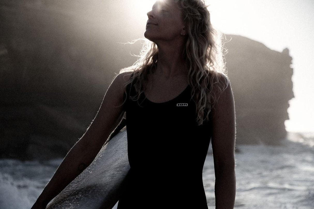 ION Monoshorty 0.5 women schwarz, Frau trägt Neoprenanzug am Strand mit Surfboard