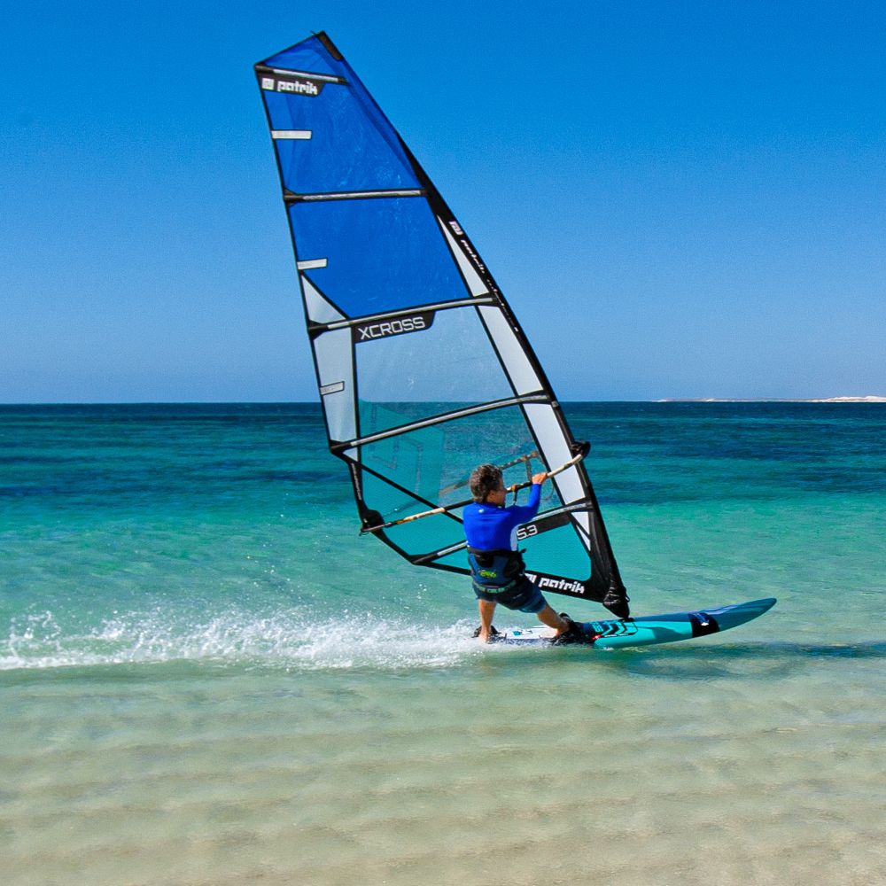 Patrik F-Cross Windsurfboard blau mit Segel am Strand, Seitenansicht im flachen Wasser