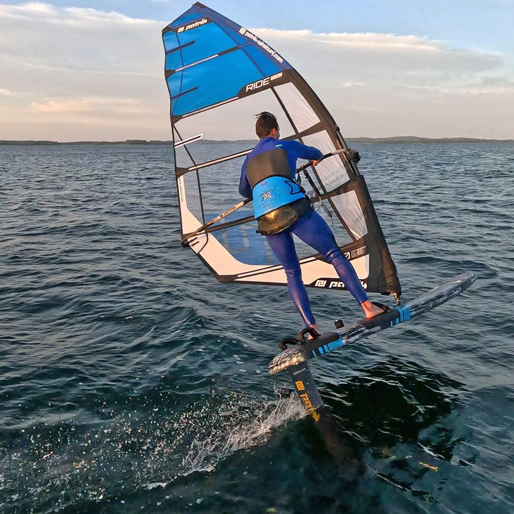 Patrik Ride Foil Windsurfboard in Blau, Fahrer in Aktion auf dem Wasser mit Segel und Foil, Seitenansicht