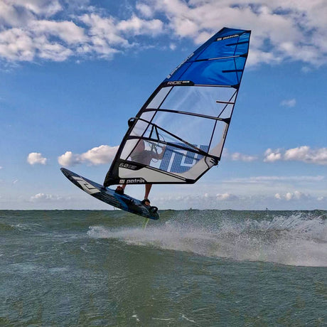Patrik Foil Ride Windsurfboard blau-schwarz in Aktion auf dem Wasser, seitliche Ansicht mit Segel und Boom