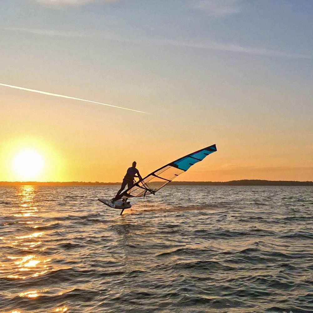 Patrik Ride Foil Windsurfboard mit blauem Segel, Athlet beim Windsurfen auf dem Wasser bei Sonnenuntergang