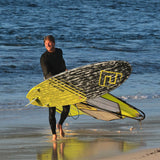 Patrik F-Wave Wellenreiter mit gelbem und schwarzem Surfboard am Strand mit Welle im Hintergrund