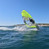 Patrik F-Wave Windsurfboard mit neongelber und grauer Segel beim Wellenreiten im Meer, Seitenansicht
