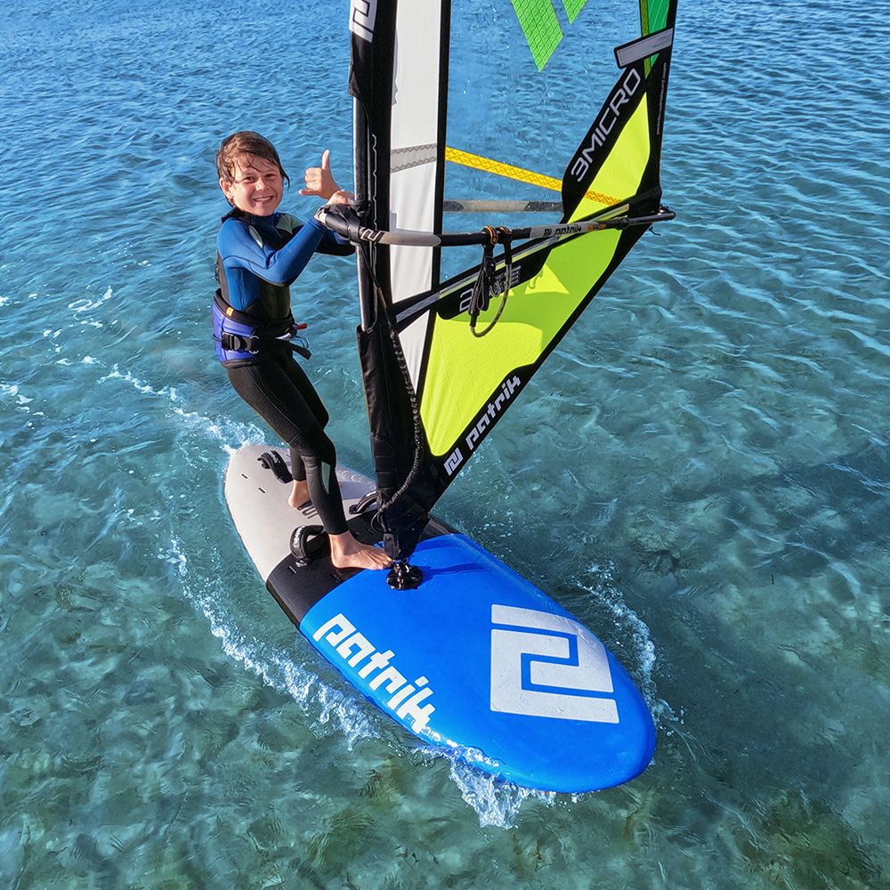 Patrik Micro Ride Windsurf-Board blau mit neongelb-grünem Segel, Kind beim Windsurfen im Wasser