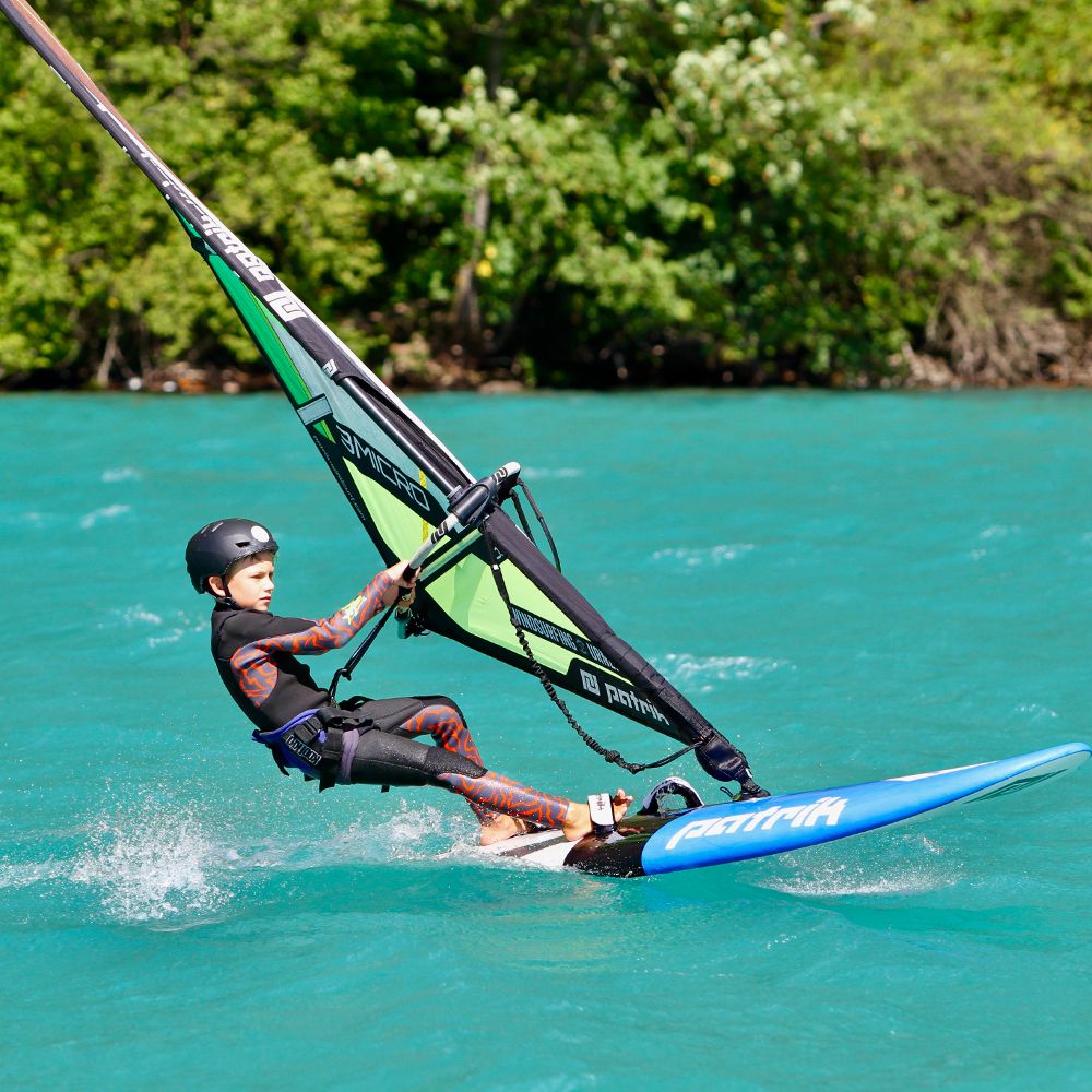 Patrik Micro Ride Windsurfboard mit grünem Segel, Kind beim Windsurfen auf türkisfarbenem Wasser in Aktion