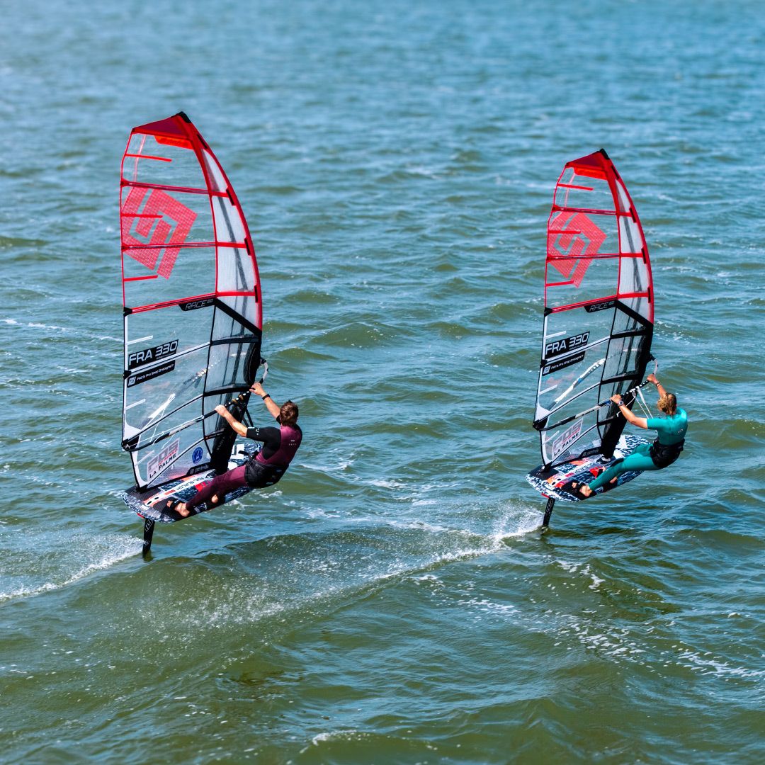 Patrik Race HA Gen2 Windsurfboard rot-weiß mit rotem Segel, zwei Windsurfer im Wasser in Frontalansicht