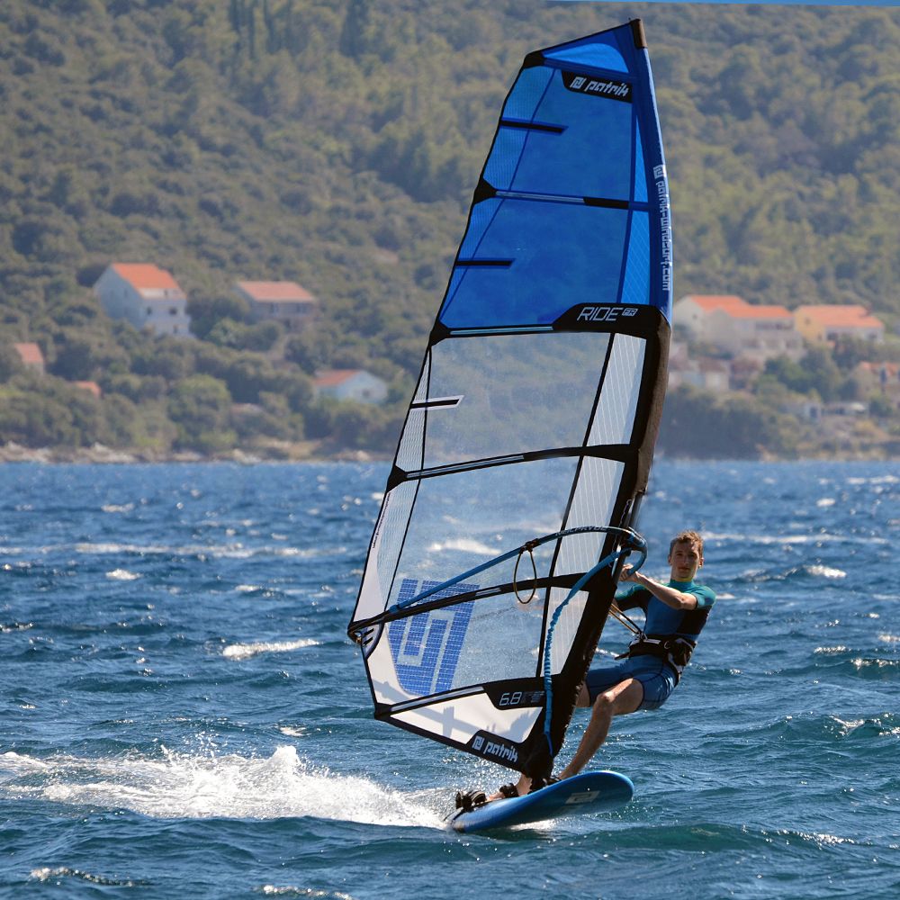 Patrik Ride FR Windsurfboard blau mit schwarzem Rahmen, Seitenansicht auf dem Wasser mit Windsurfer