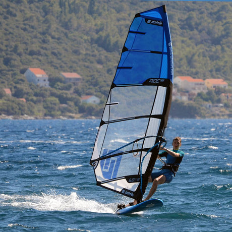 Patrik Ride FR Windsurfboard blau mit schwarzem Rahmen, Seitenansicht auf dem Wasser mit Windsurfer
