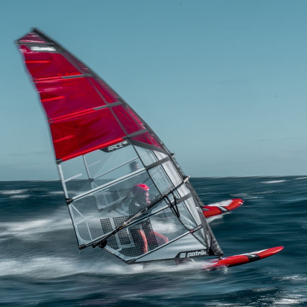 Patrik Slalom Windsurfboard mit rotem Segel und grauem Rumpf, Frontansicht im Wasser