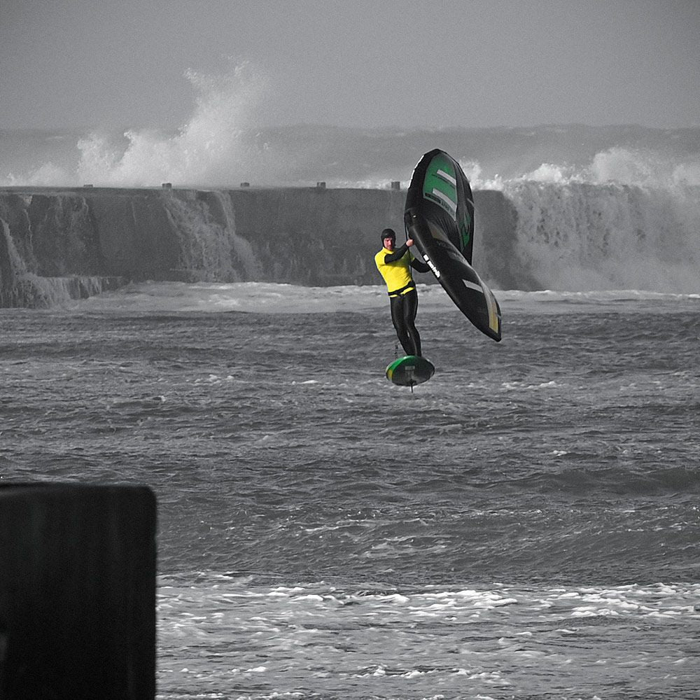 Patrik Wing V2 Volant Cross Kite in Schwarz-Grün, in Aktion über stürmischer See mit Kitesurfer in gelbem Neoprenanzug