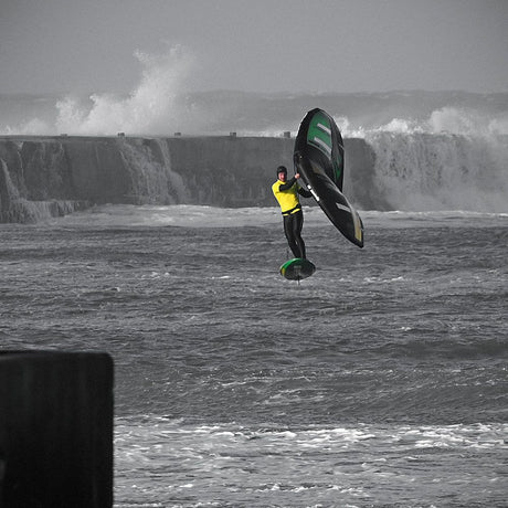 Patrik Wing V2 Volant Cross Kite in Schwarz-Grün, in Aktion über stürmischer See mit Kitesurfer in gelbem Neoprenanzug