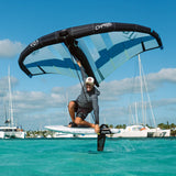 Chase WB-X Foilboard schwarz-blau, Athlet in Aktion über türkisem Wasser mit Foil, Segelboote im Hintergrund